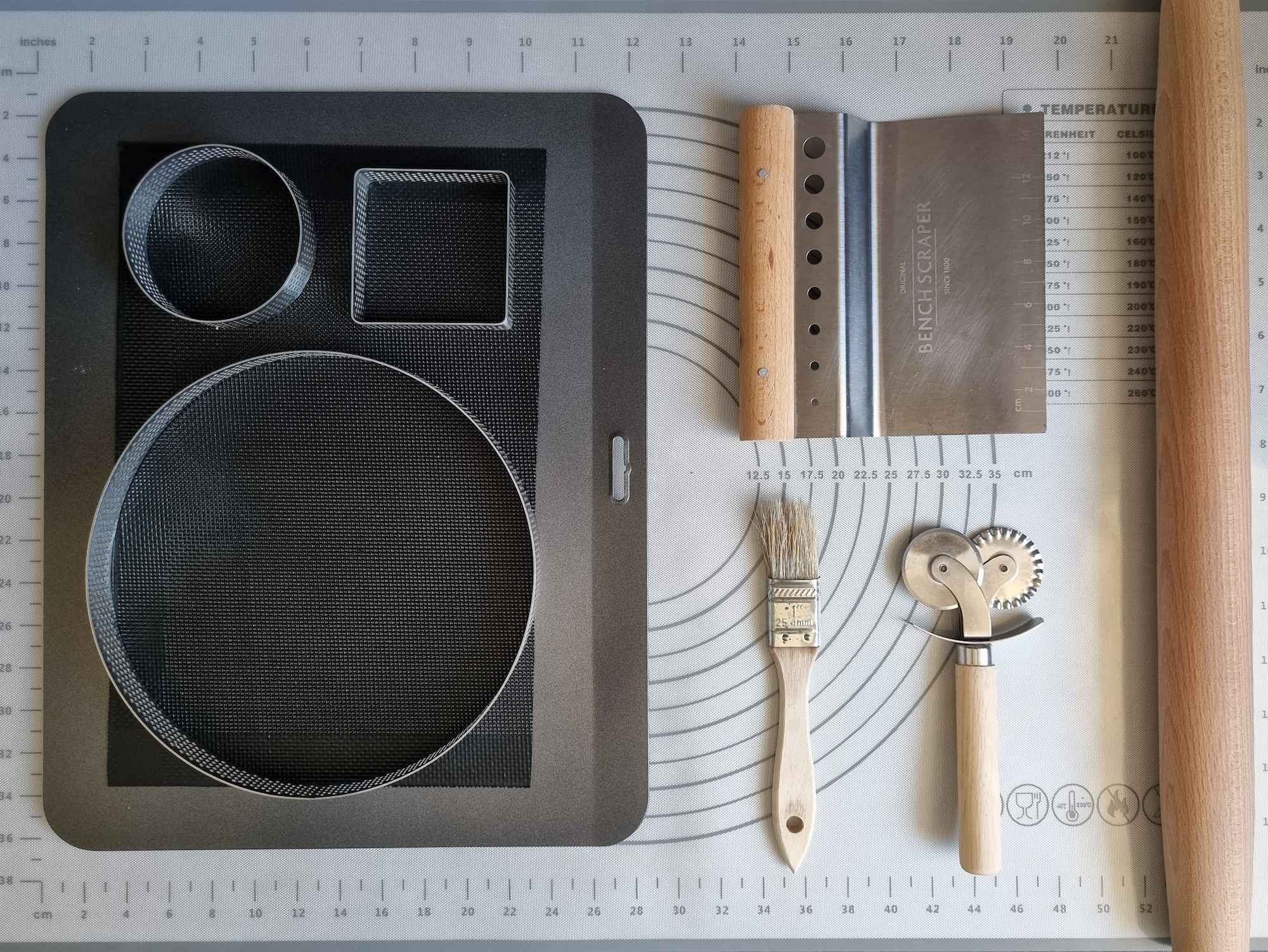 Organised kitchen equipment with a pastry mat, baking tray, baking mat, tart rings, bench scraper, brush, cutting wheel and rolling pin.