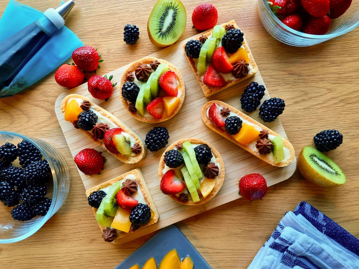 Fruit tartelettes on a wooden board, surrounded by fresh fruit and baking tools.