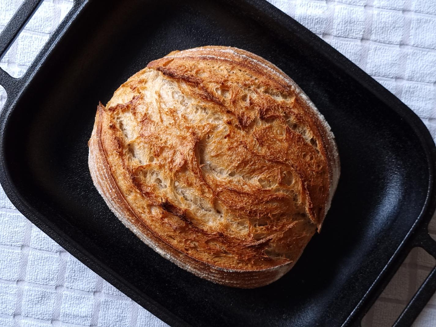 A baked, golden brown sourdough loaf, Pain De Campagne, sitting in a cast iron bread pan.